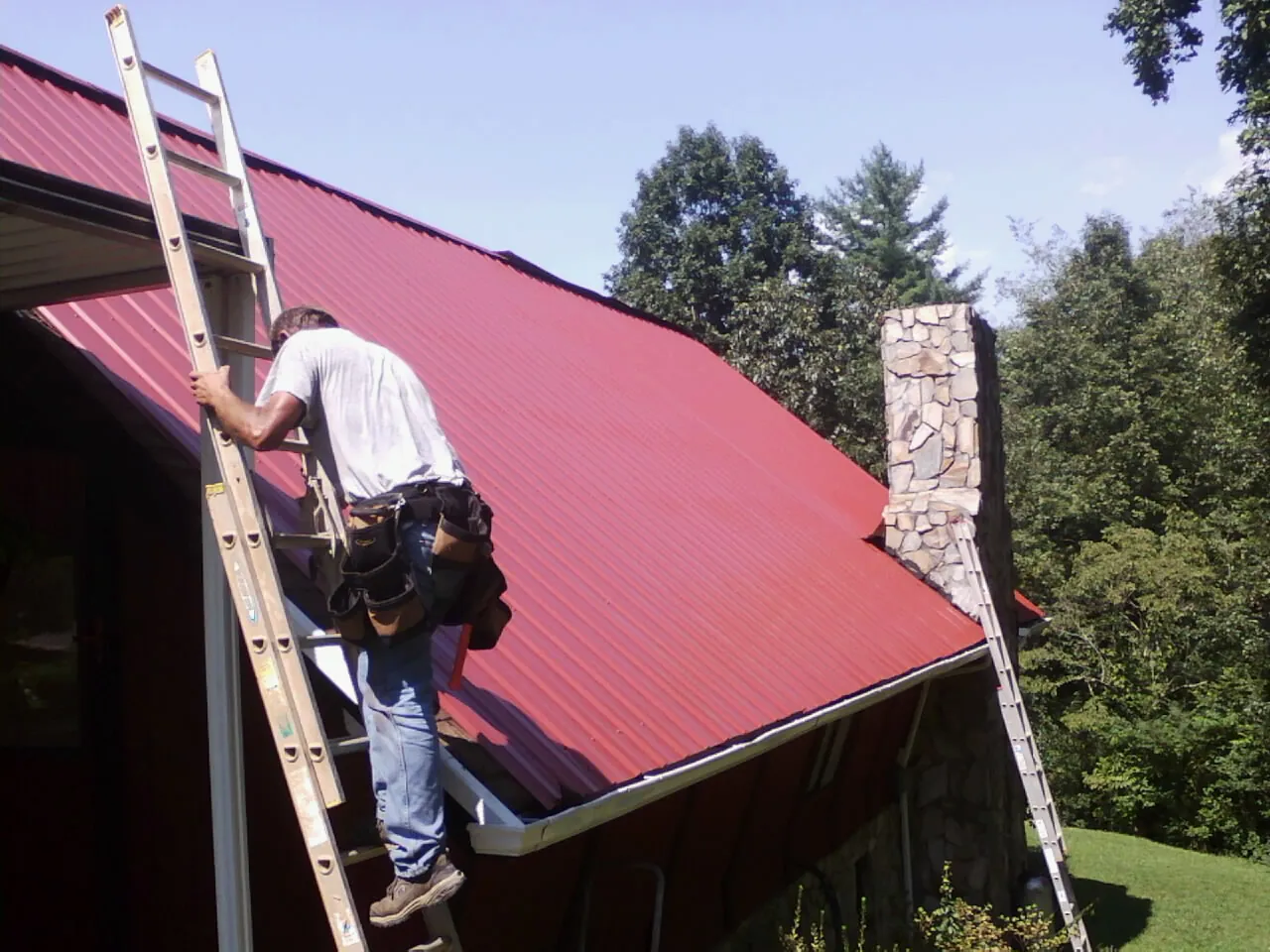 Roofer on a ladder beside a red metal roof and stone chimney — Soffit & Fascia Repair by Roofing Brothers in Soap Lake